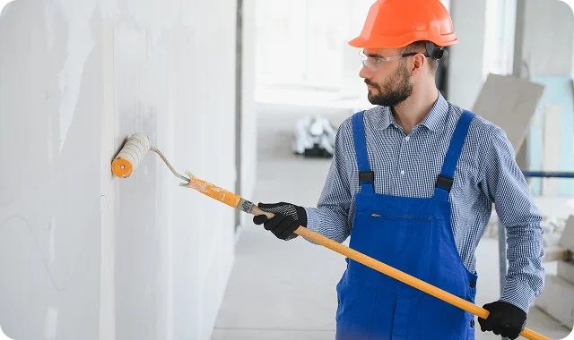 Man in blue overalls and orange hard hat painting a wall with a roller in a bright, unfinished room.